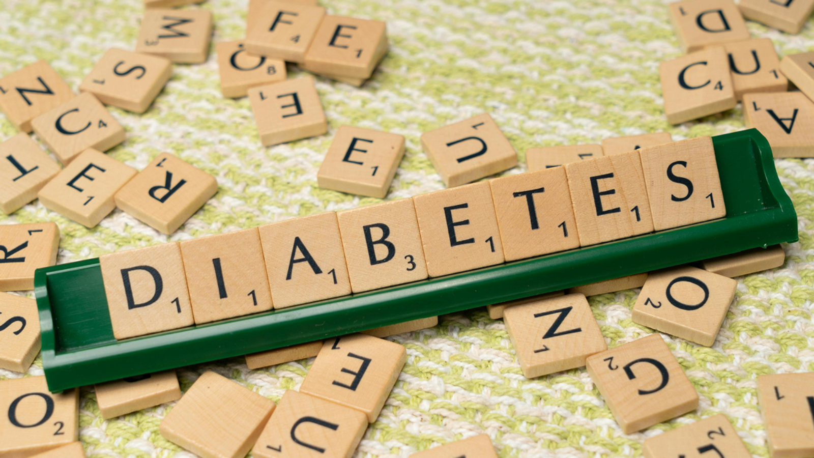 Wooden letter tiles on a green rack spelling out the word "DIABETES" against a scattered tile background.