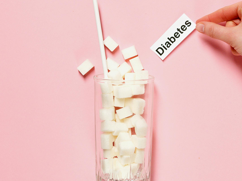 A tall glass filled to the brim with white sugar cubes on a pink background, with a hand holding a "Diabetes" label.