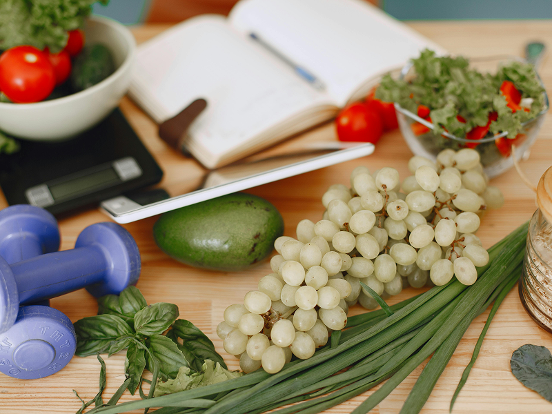 A tabletop filled with healthy food like grapes and greens, alongside a notebook, digital scale, and blue dumbbells.