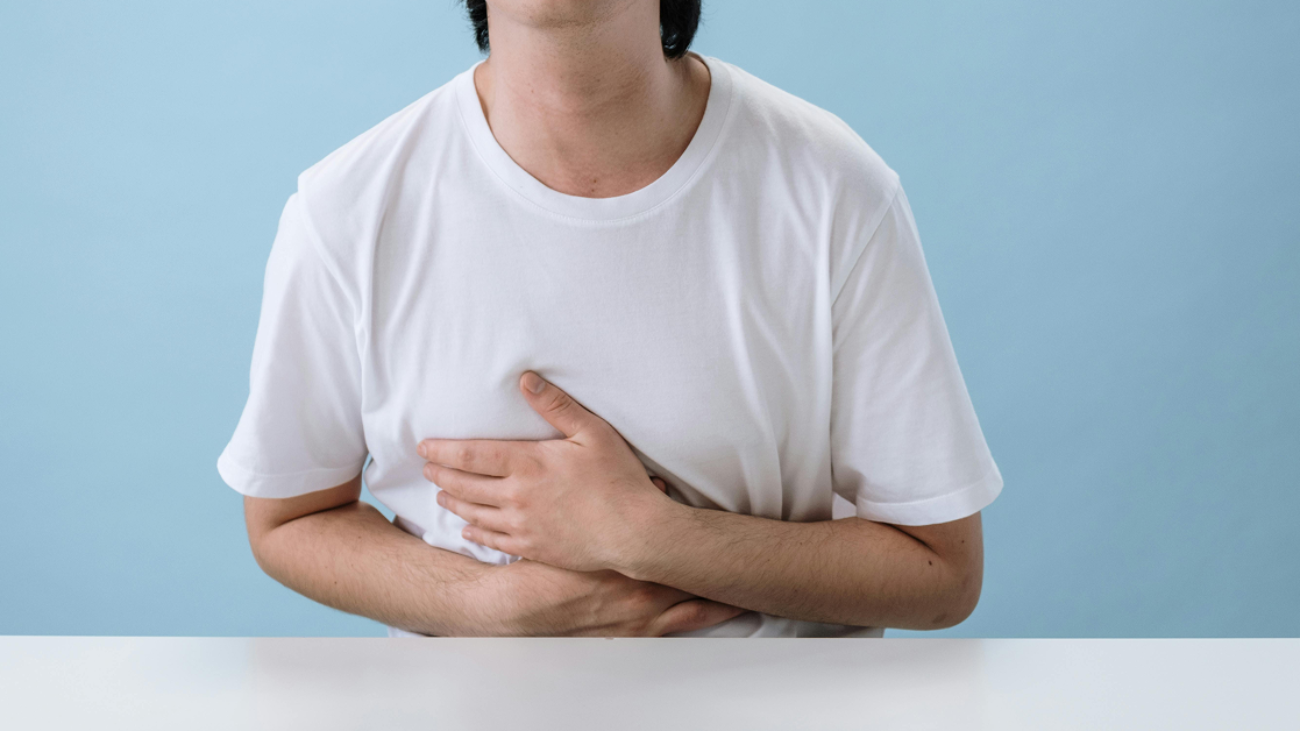 A person in a white t-shirt sitting at a table and holding their stomach in discomfort.