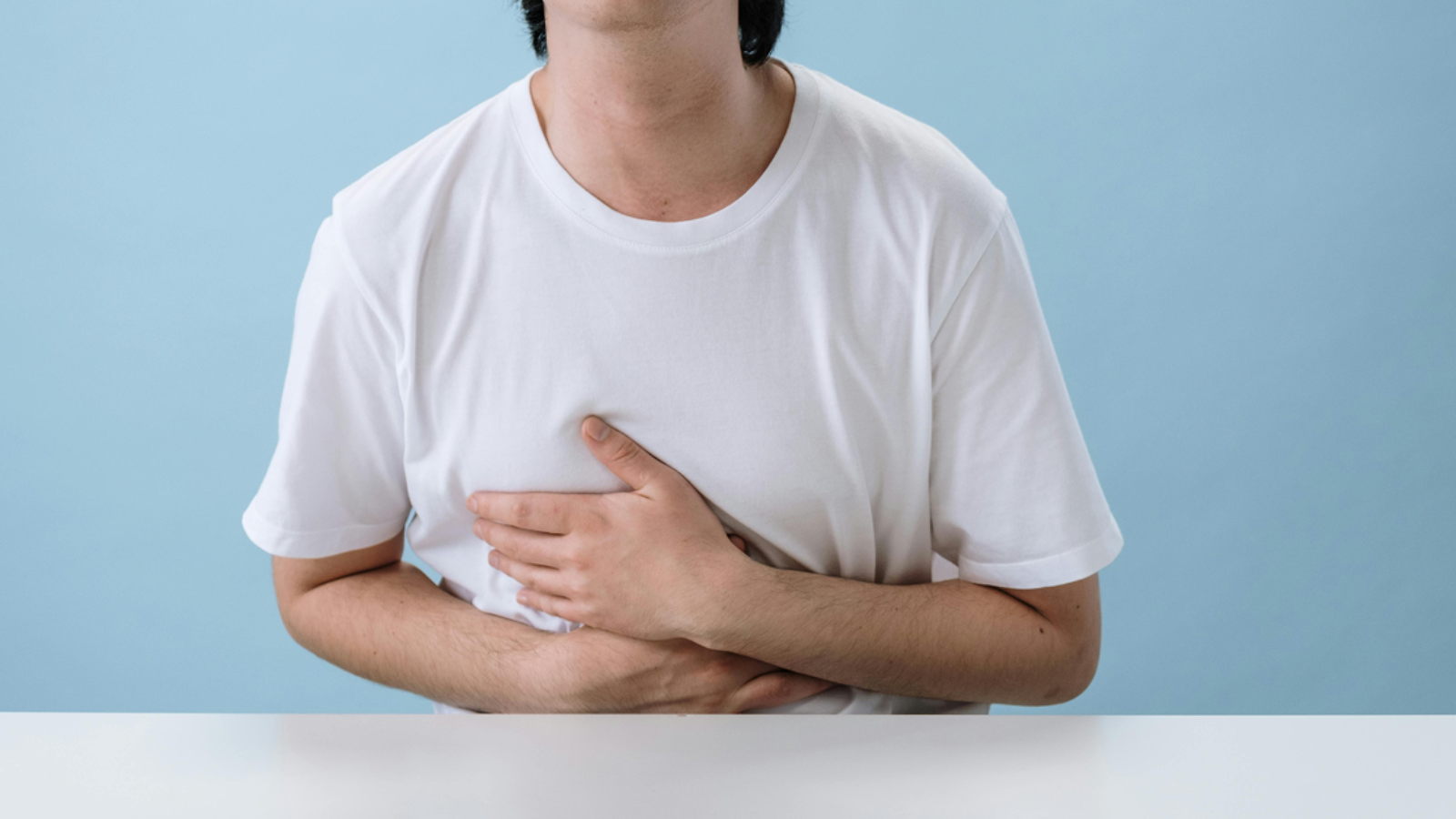 A person in a white t-shirt sitting at a table and holding their stomach in discomfort.