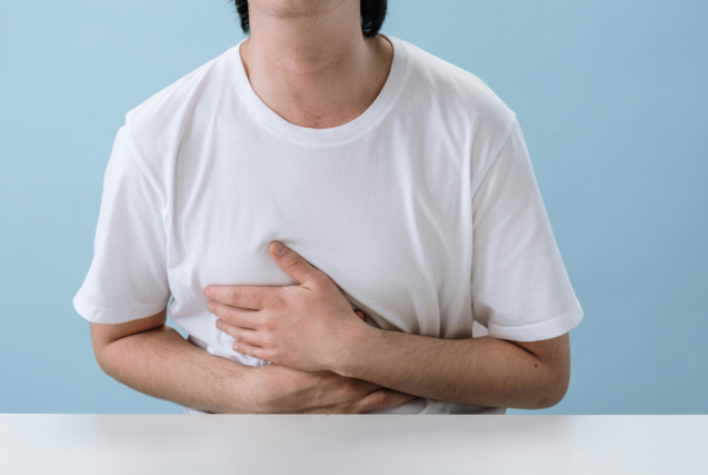 A person in a white t-shirt sitting at a table and holding their stomach in discomfort.