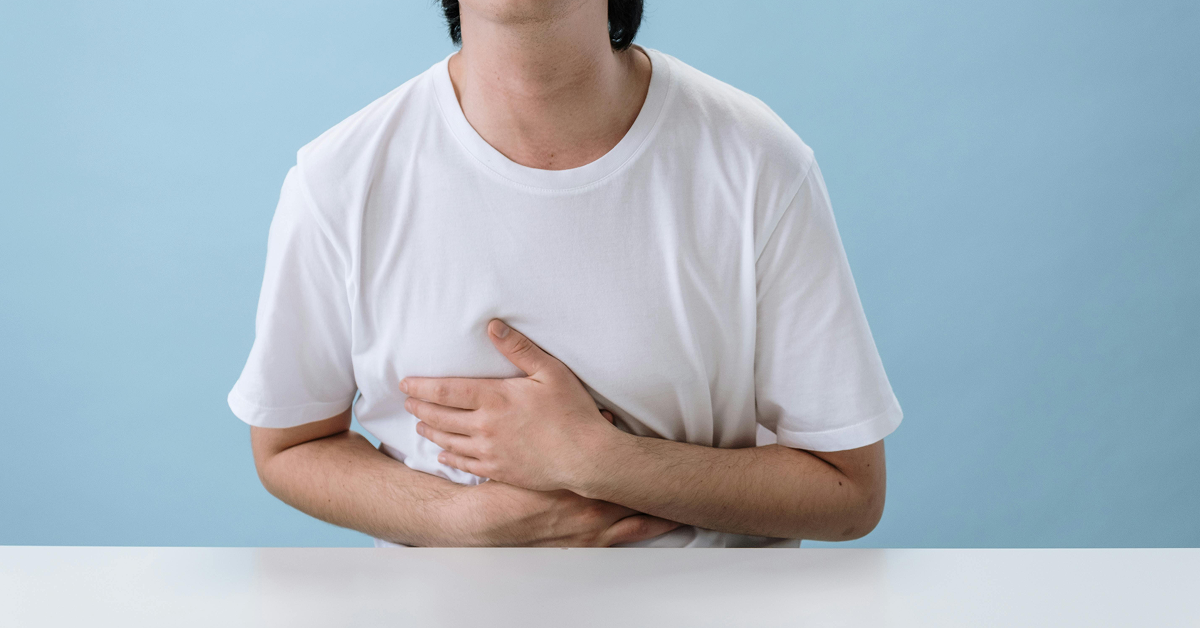 A person in a white t-shirt sitting at a table and holding their stomach in discomfort.