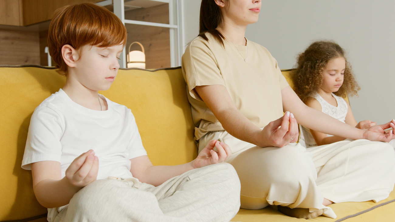 A woman and two children sitting on a yellow sofa with their eyes closed in a meditation pose.
