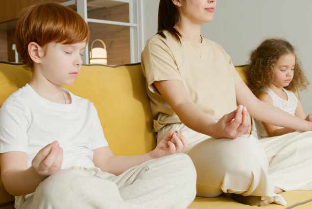 A woman and two children sitting on a yellow sofa with their eyes closed in a meditation pose.