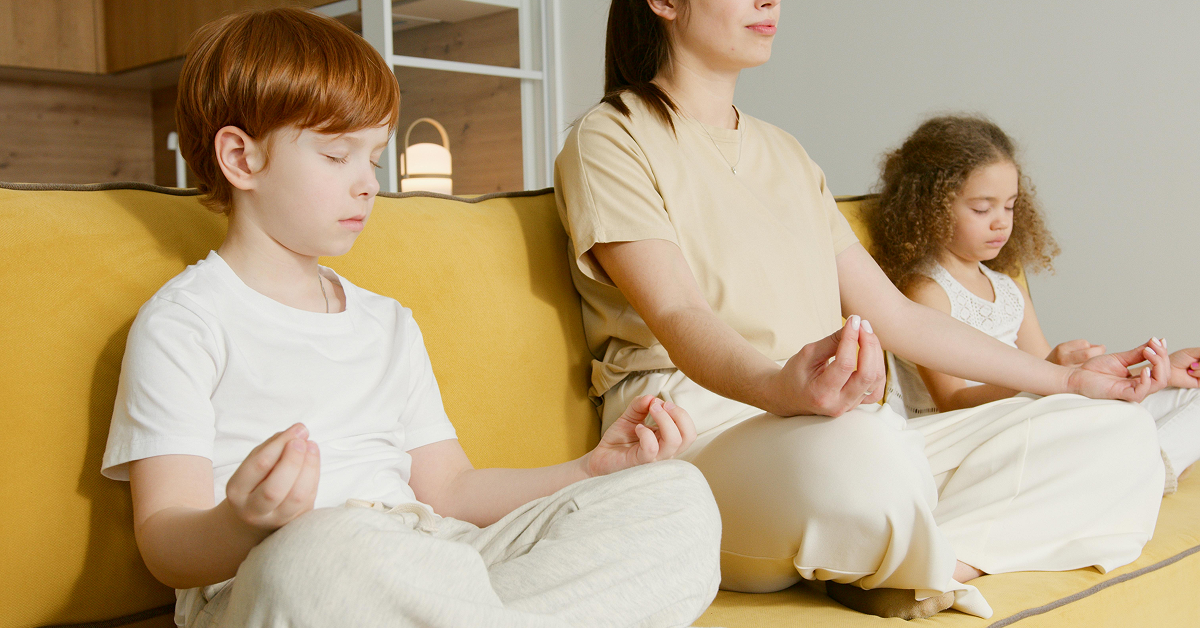 A woman and two children sitting on a yellow sofa with their eyes closed in a meditation pose.