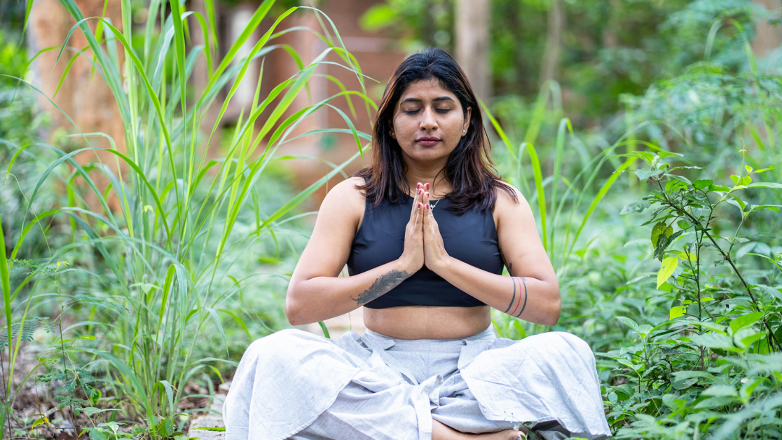 A woman sitting in a cross-legged meditation pose with her hands in prayer position, surrounded by lush green foliage.