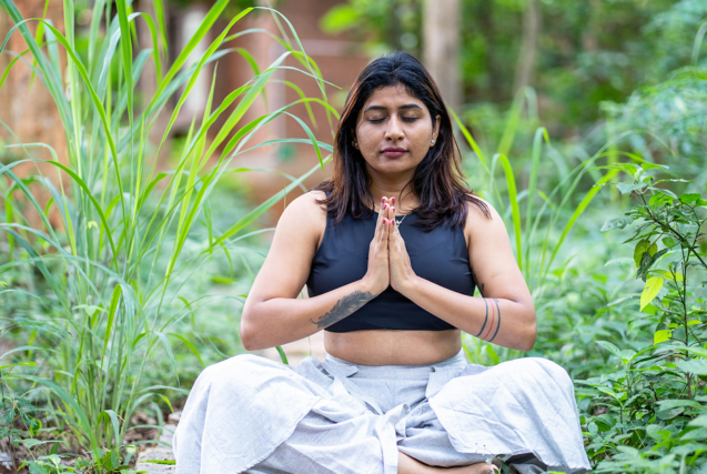 A woman sitting in a cross-legged meditation pose with her hands in prayer position, surrounded by lush green foliage.