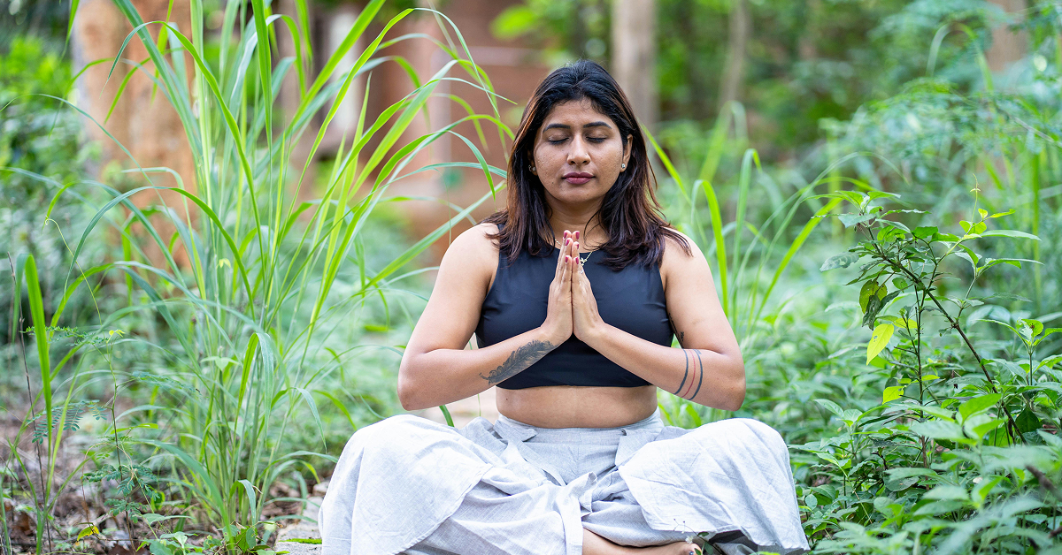 A woman sitting in a cross-legged meditation pose with her hands in prayer position, surrounded by lush green foliage.