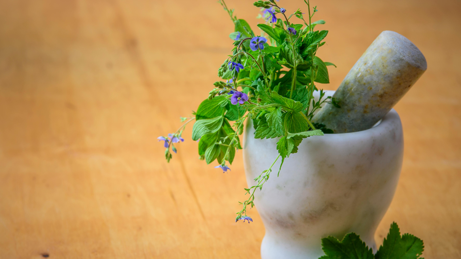 A white marble mortar and pestle filled with fresh green herbs and small purple flowers on a wooden surface.