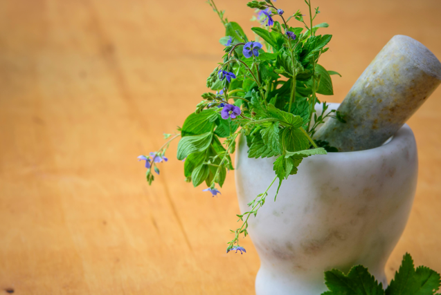 A white marble mortar and pestle filled with fresh green herbs and small purple flowers on a wooden surface.