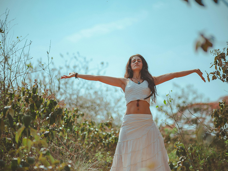 A woman in white clothing standing in a field with her arms outstretched and eyes closed toward the sky.