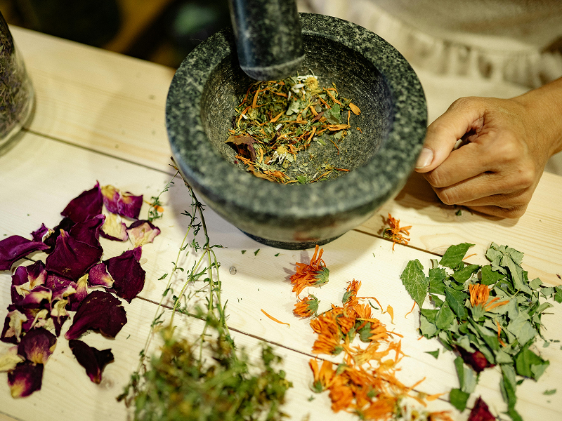 A person using a stone mortar and pestle to grind a mixture of dried flowers and herbs on a light wooden table.