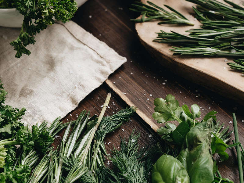 A variety of fresh green herbs, including rosemary, parsley, and basil, scattered on a dark wooden surface with grains of salt.