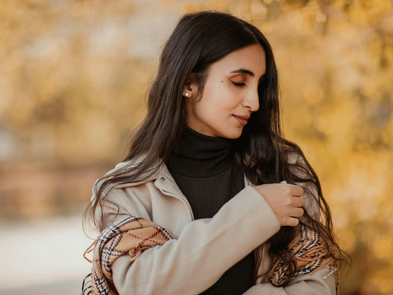 A woman in a beige coat looking down thoughtfully while running her hand through her long, dark hair.