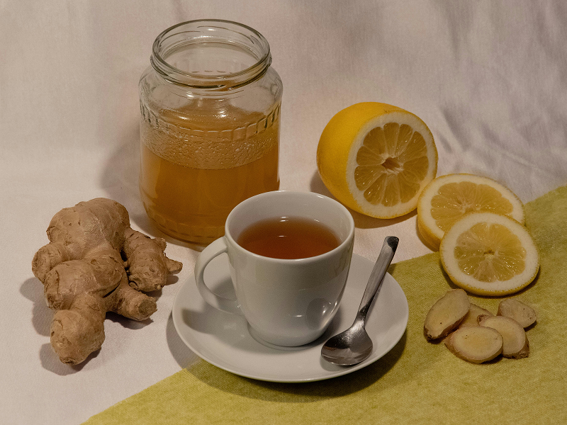 A white cup of tea on a saucer with a spoon, surrounded by a jar of honey, fresh ginger root, and lemon slices.
