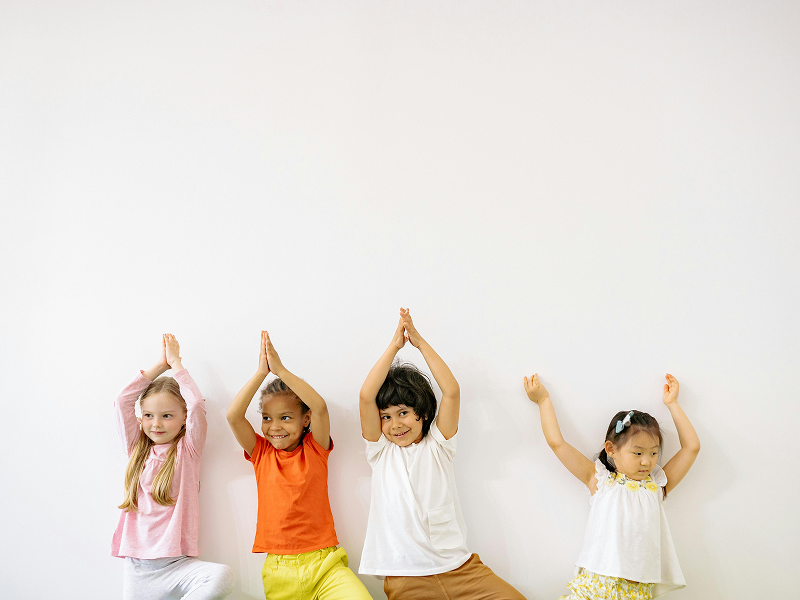 Four young children standing against a white wall practicing yoga poses with their arms raised.
